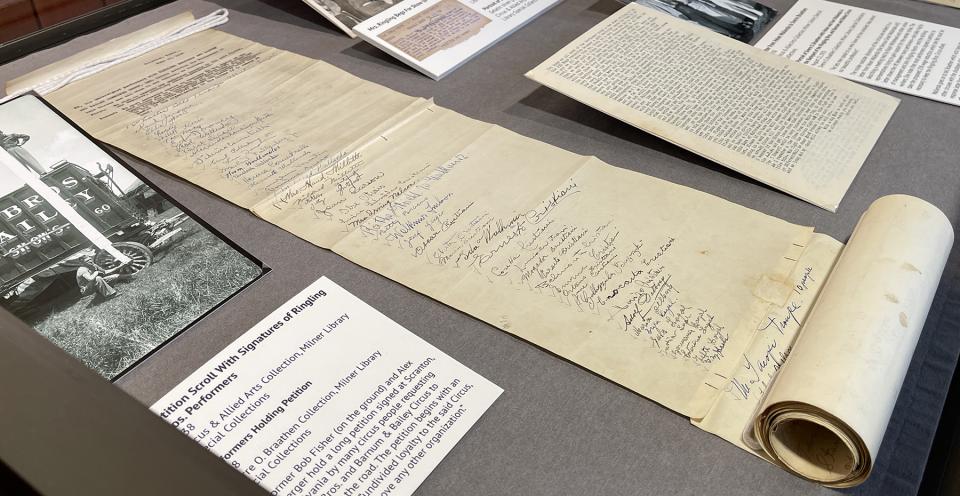 Closeup photograph showing a display case with a photo of a man standing atop a circus wagon holding a scroll that extends to the ground while another man crouches holding the bottom of the page. A partially unrolled paper scroll (pictured in the other image) with signatures is placed alongside the photograph.