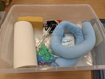 Items within ISU mini disaster kits including pig socks; flashlights and batteries; plastic sheeting; tape and scissors; personal protective equipment - plastic apron, gloves, mask; paper towel. All in a large plastic tub.